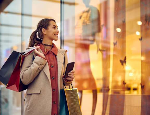 A woman is standing outside a store holding shopping bags and a phone.