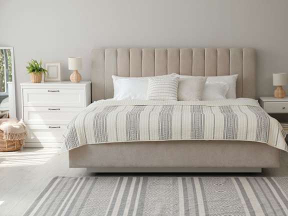 A bed with a striped comforter and two pillows sits in a room with a white nightstand and lamp beside it.