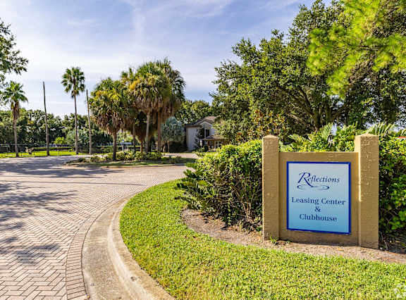 A sign for Reflections Leasing Center & Clubhouse is in front of a brick path and greenery.