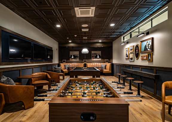 A foosball table is in the middle of a room with leather chairs and a bar area.