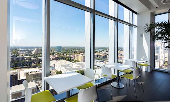A room with white tables and chairs with a view of the city outside the windows.