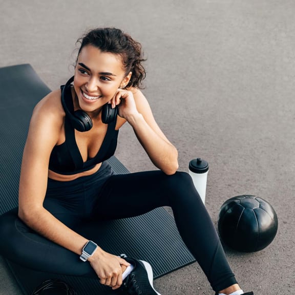 a woman sitting on a fitness mat with headphones on and a medicine ball next to her