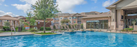 a swimming pool with a tree and lounge chairs in front of a building at Ovation at Lewisville Apartments, Texas , 75067