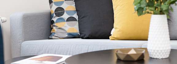 A grey couch with a yellow pillow and a vase with white flowers on a table.