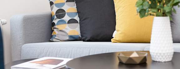 A grey couch with a yellow pillow and a vase with white flowers on a table.