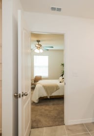 A bathroom with a dark vanity opens to a bedroom with a white bed and ceiling fan.