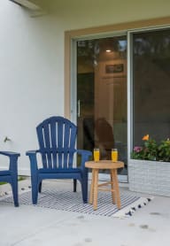 A cozy patio features two blue Adirondack chairs, a small table with drinks, and a flower planter by a sliding glass door.