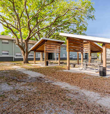 A wooden pavilion with a white roof is surrounded by a gravel area and a tree with green leaves.