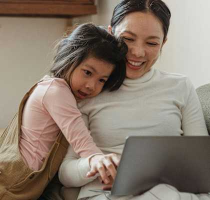 a little girl hugging her mother who is sitting on a couch looking at a laptop computer