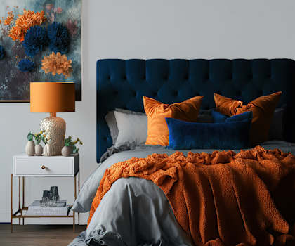 A bed with a blue headboard and orange bedding is in the foreground with a lamp and painting in the background.