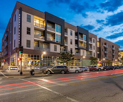 A modern apartment building is shown at dusk with cars parked in front.