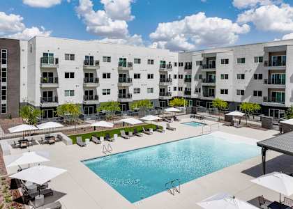 A large swimming pool surrounded by lounge chairs and umbrellas in front of a white apartment building.