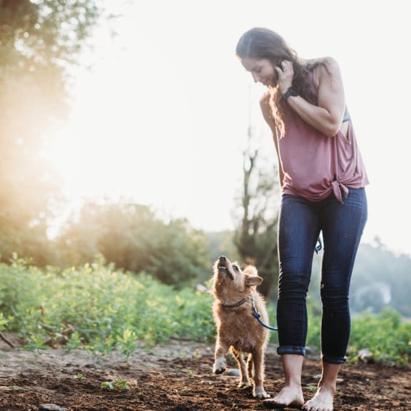 A woman and her dog are standing on a dirt path in a field.