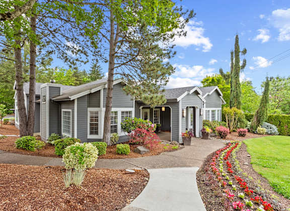 A house with a grey roof and white trim is surrounded by a garden with red flowers and green shrubs.