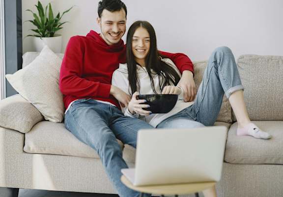 a man and a woman sitting on a couch eating popcorn and watching TV on a laptop