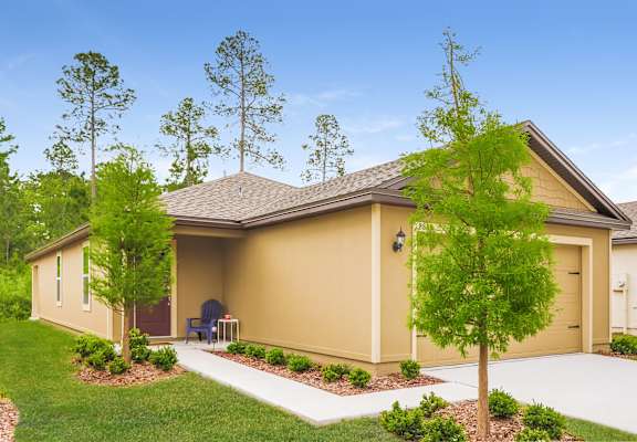 A single-story beige house with a brown garage door features a blue chair on the porch under a partly cloudy sky.