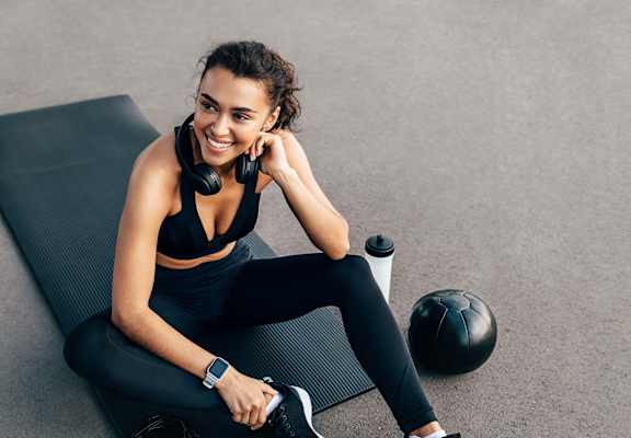 A woman in workout gear sits on a mat with a water bottle, a soccer ball, and headphones.