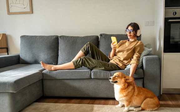 A woman is sitting on a grey couch with her legs crossed, holding a yellow object in her hand, and a dog is sitting next to her.