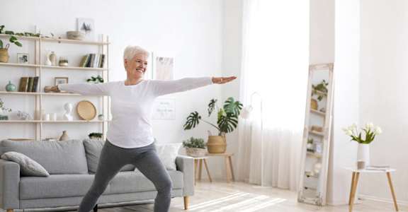 A woman is practicing yoga in a bright room with a grey couch and a wooden floor.