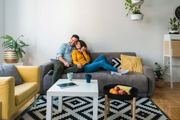 A couple is sitting on a grey couch in a living room with a yellow chair and a coffee table.