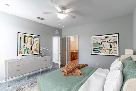 A bedroom with a bed, a ceiling fan, and two paintings on the wall.
