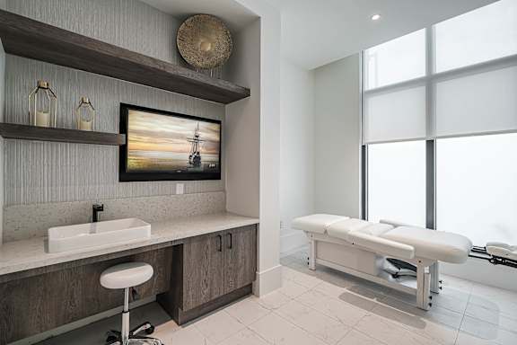 A medical examination table is in a room with a sink and a picture of a sunset.