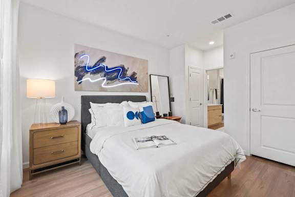 A bedroom with a large bed, a painting above it, and a wooden nightstand with a lamp.