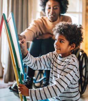 a boy in a wheelchair paints with a paintbrush with his mother in the background