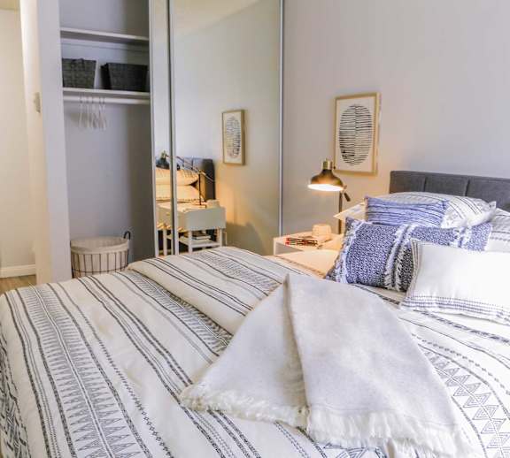 A bed with blue and white striped bedding is in the foreground of a room with a mirrored closet and a lamp.