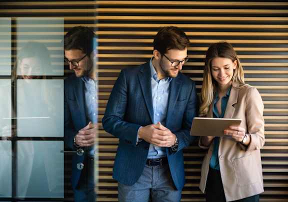 Two people in business attire are looking at a tablet while standing in front of a window with blinds.