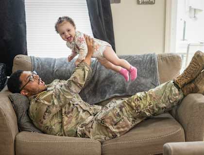 A man in military uniform is lying on a couch with a baby on his feet.