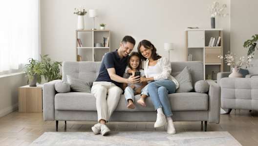 A family of three is sitting on a grey couch in a well-lit room with a large window.