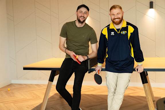 Two men are standing in a room with a table and holding paddles and a table tennis ball.