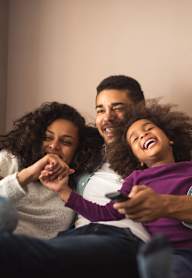 Three people are sitting on a yellow couch, laughing and looking at a phone.