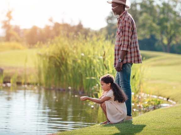 A man and a girl are standing by a pond on a sunny day.