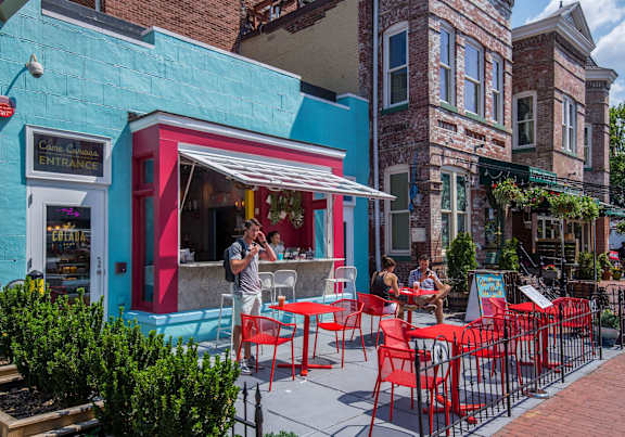 A man is standing outside a restaurant with red chairs and tables.