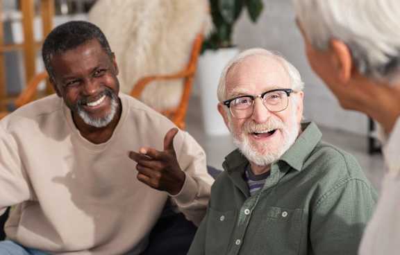 Two men are having a conversation in a room with a white furry chair in the background.