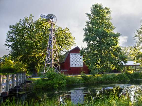A windmill stands next to a red barn by a body of water.