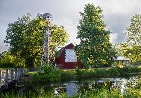 A windmill stands next to a red barn by a body of water.