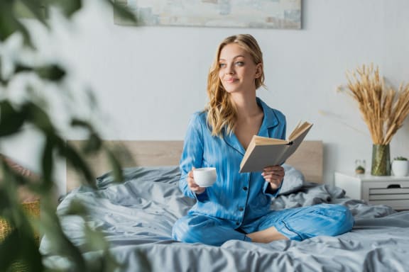 a woman sitting on her bed reading a book and holding a cup of coffee