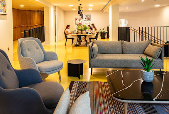 A living room with a grey couch, a grey armchair, a black coffee table and a woman sitting at a table in the background.