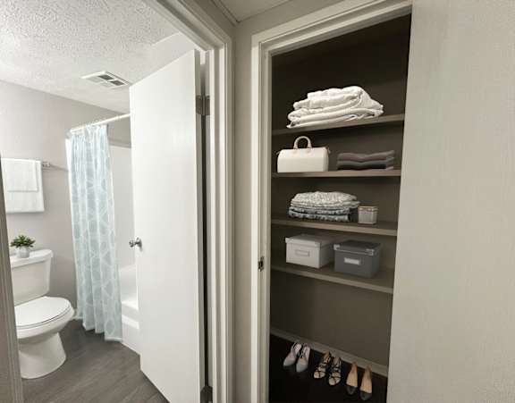 A bathroom with a toilet, a shower curtain, and a shelf with towels and shoes.