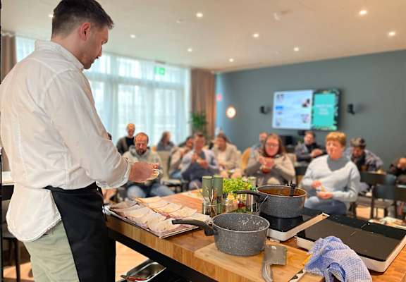 A man in a white shirt and black apron is cooking in front of a group of people.