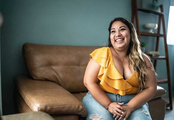 A woman in a yellow top and blue jeans sits on a brown leather couch.