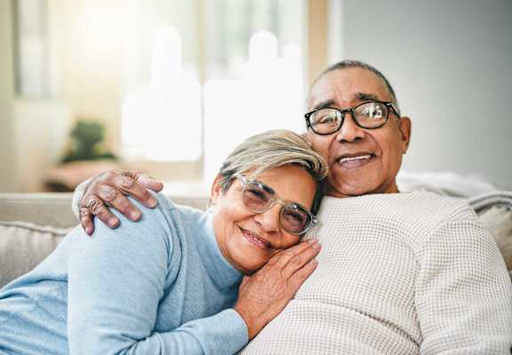 An older couple is sitting on a couch, smiling and embracing each other.