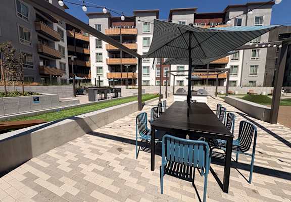 A black table with blue chairs and a black umbrella in front of apartment buildings.