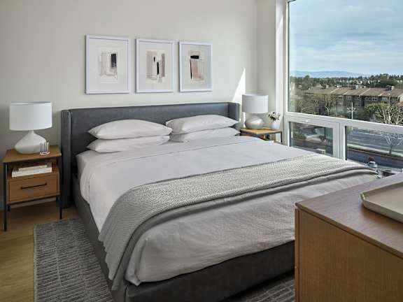 A bedroom with a large bed, a nightstand with a lamp, and a window overlooking a cityscape.