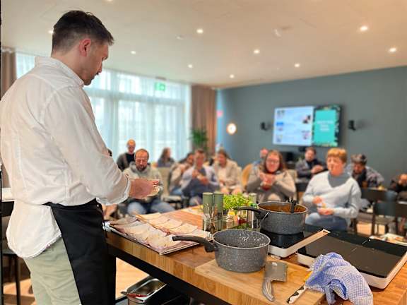 A man in a white shirt and black apron is cooking in front of a group of people.