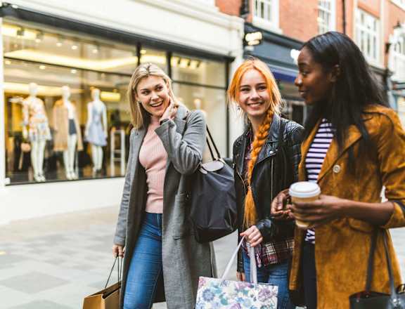 Three women are standing outside a store, smiling and holding shopping bags.