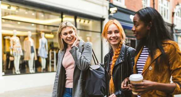 Three women are standing outside a store, smiling and holding shopping bags.